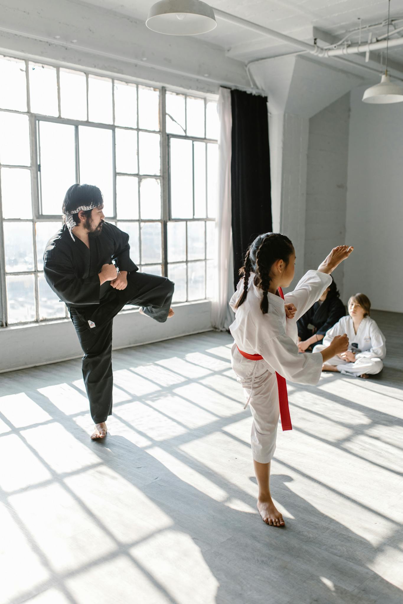 A martial arts class with a girl and an adult practicing kicks in a sunlit indoor space.