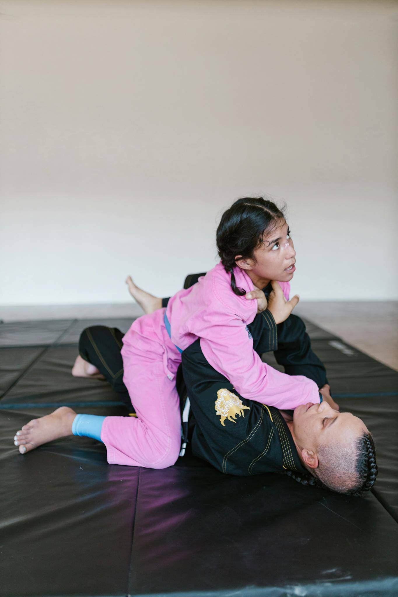Two kids in martial arts uniforms practicing grappling on a gym mat indoors.